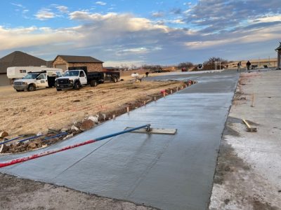 A concrete driveway is being poured in a residential area, with workers and equipment visible on the site.
