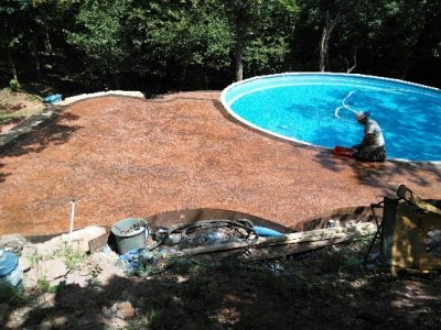 A man is working on maintaining a pool in a backyard, surrounded by grass and trees.
